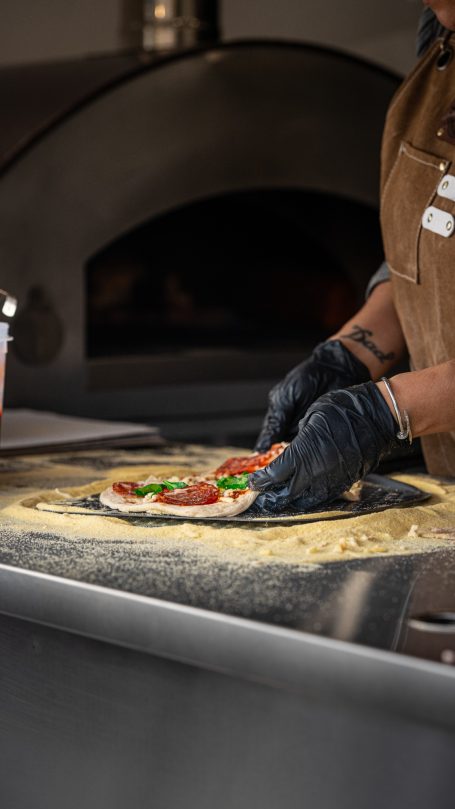 Person preparing a pizza with toppings on a dough in a kitchen setting.