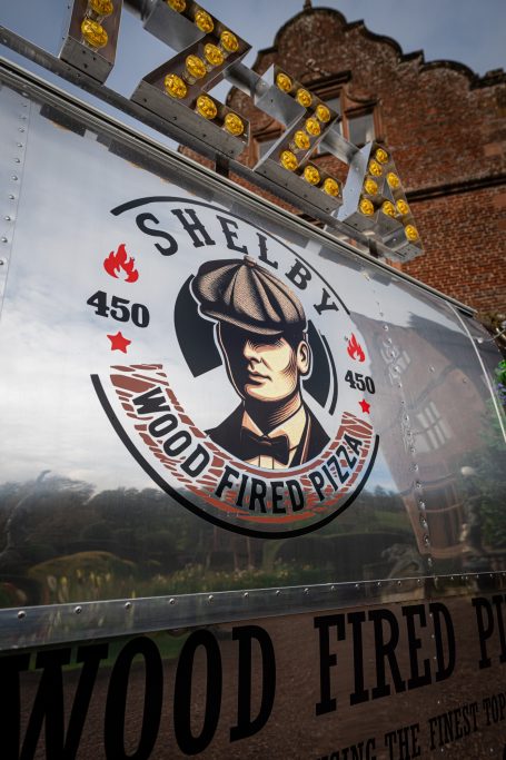Sign for "Shelby Wood Fired Pizza" featuring a man in a flat cap and pizza graphics.