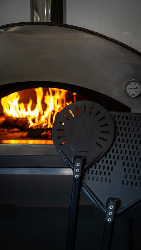 Firewood pizza oven with flames and a black peel tool in the foreground.