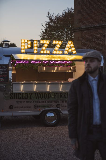 A man stands near a food truck with a bright "PIZZA" sign in lights at dusk.