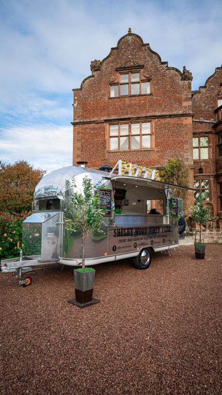 A vintage silver pizza food trailer with green foliage, set against a historic brick building.