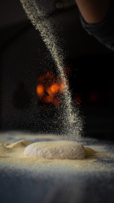 Flour being sprinkled over pizza dough with a pizza oven burning in the background.