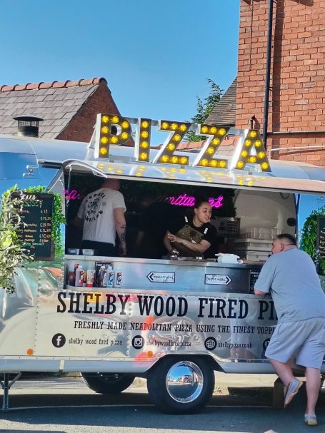 Wood-fired pizza truck with staff preparing food, vibrant signage serving a customer.