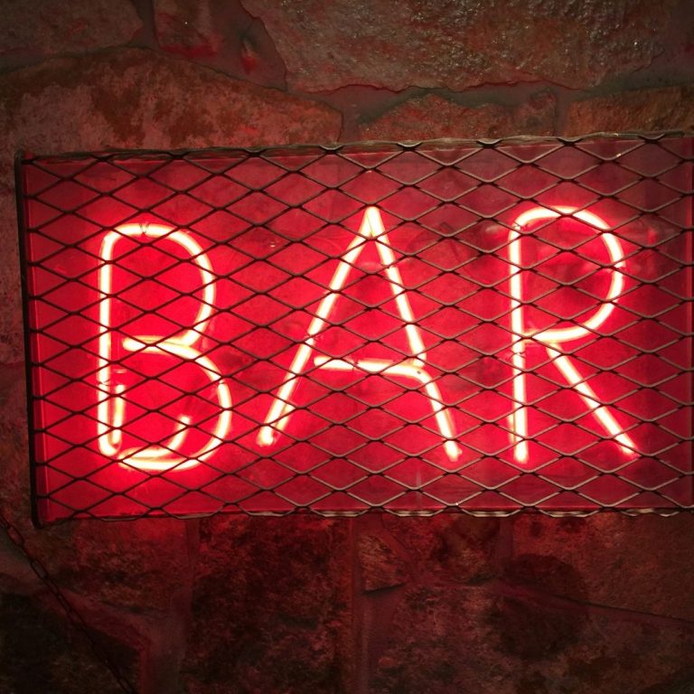 Neon sign reading "BAR" enclosed in a metal mesh frame against a stone wall.