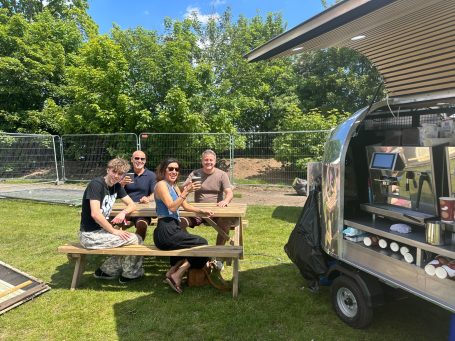 Group of four people seated at a picnic table beside a mobile coffee shop tuk-tuk in a park setting.