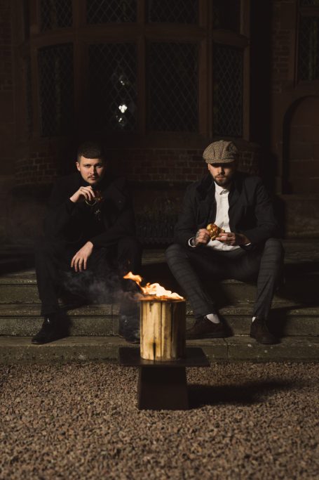 Two men sitting on steps by a small fire, one holding food and the other smoking.